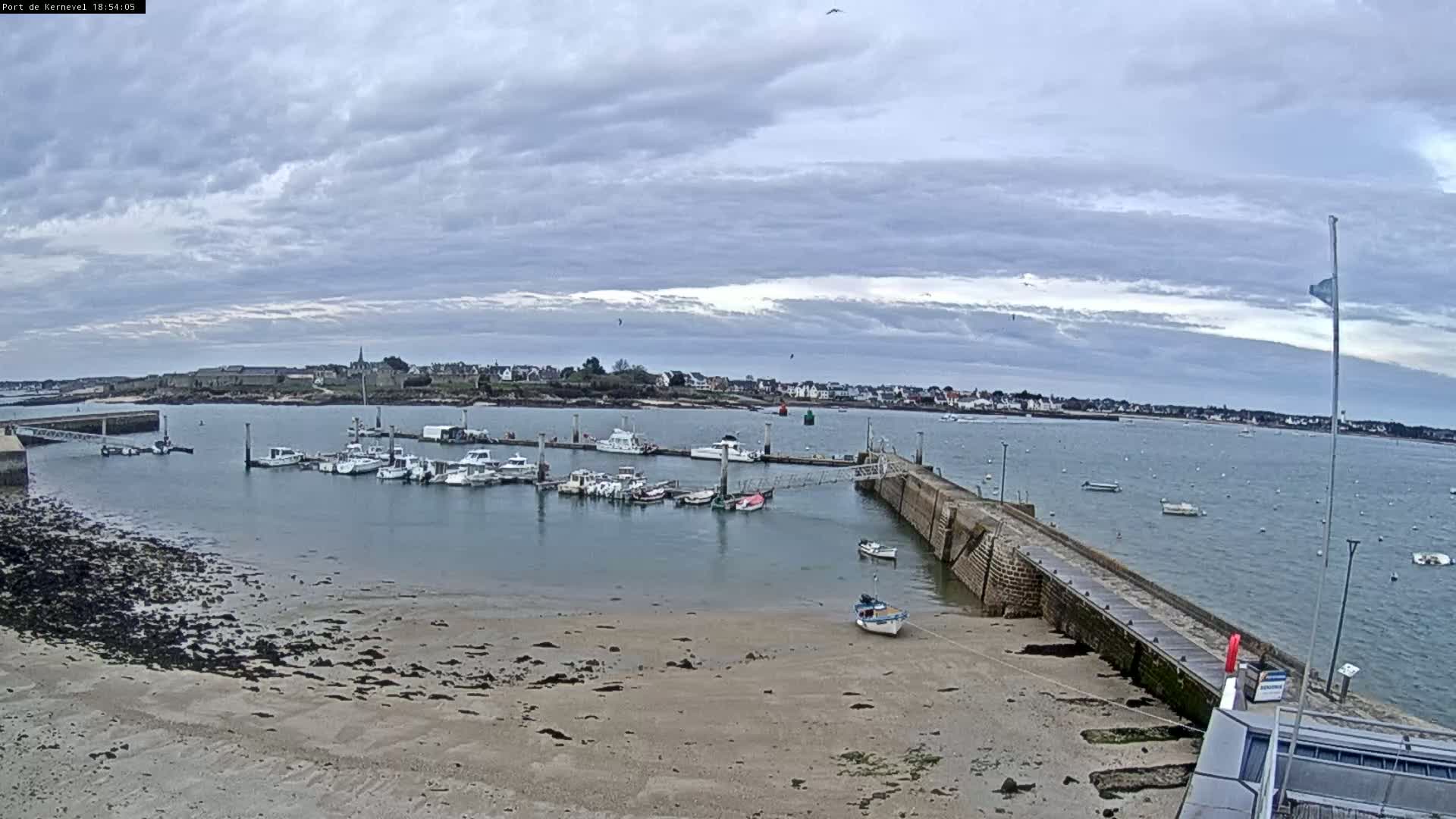 A small harbor filled with numerous boats is situated next to a sandy beach under a cloudy sky.