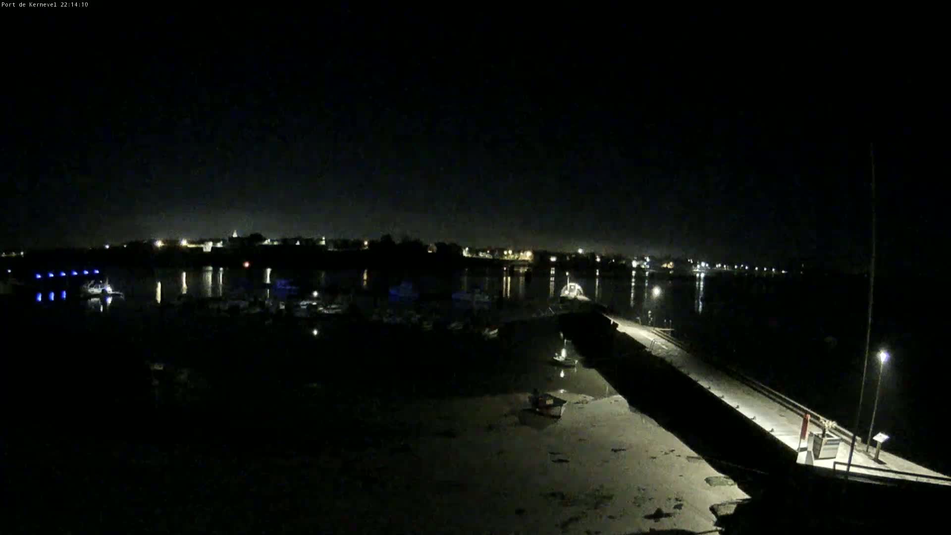 A nighttime view of a harbor with boats moored, a long pier extending into the water, and a town's lights visible in the distance.