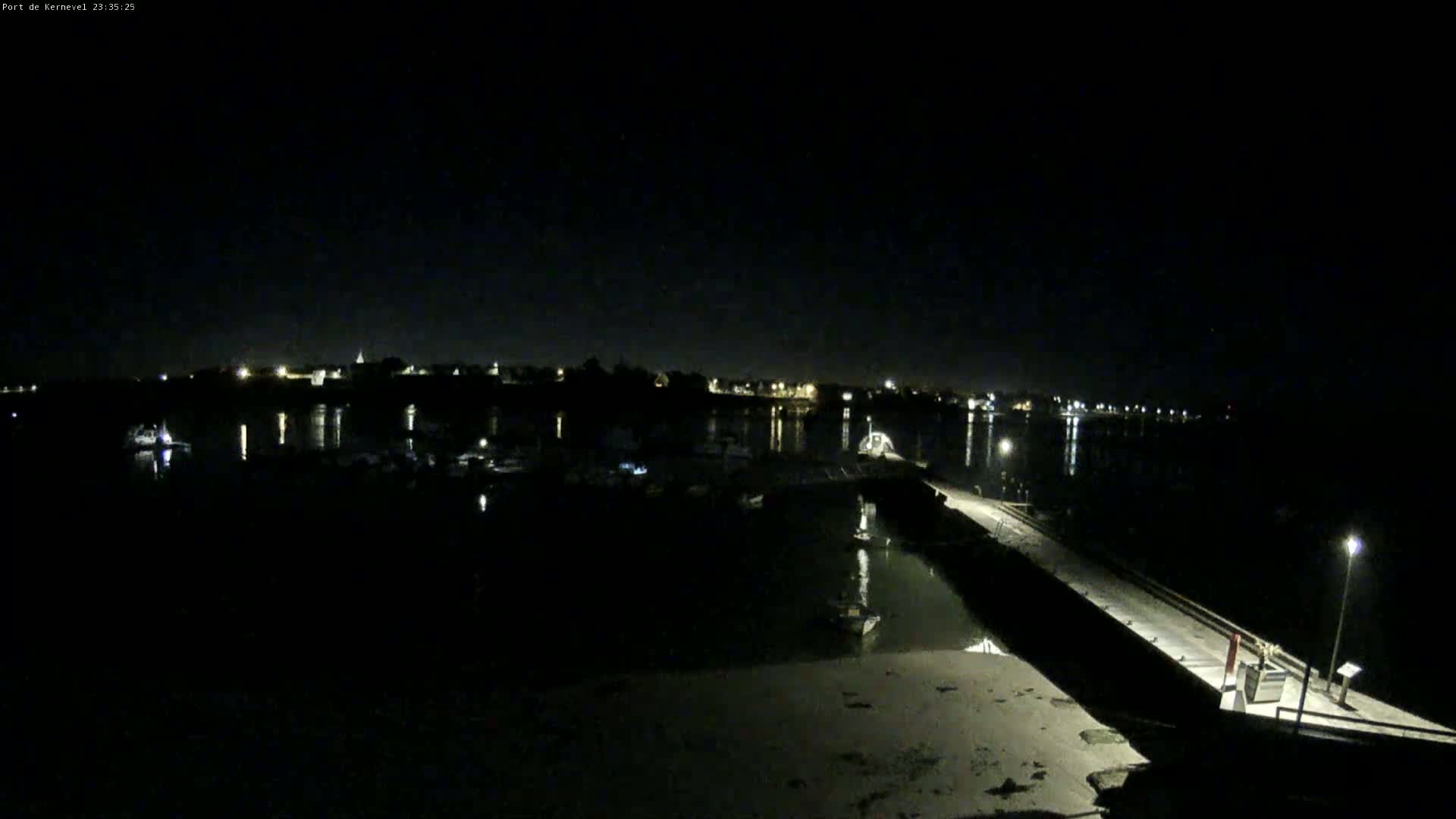 A nighttime view of a calm harbor with boats moored, illuminated by dim lights from a nearby town under a clear, dark sky.