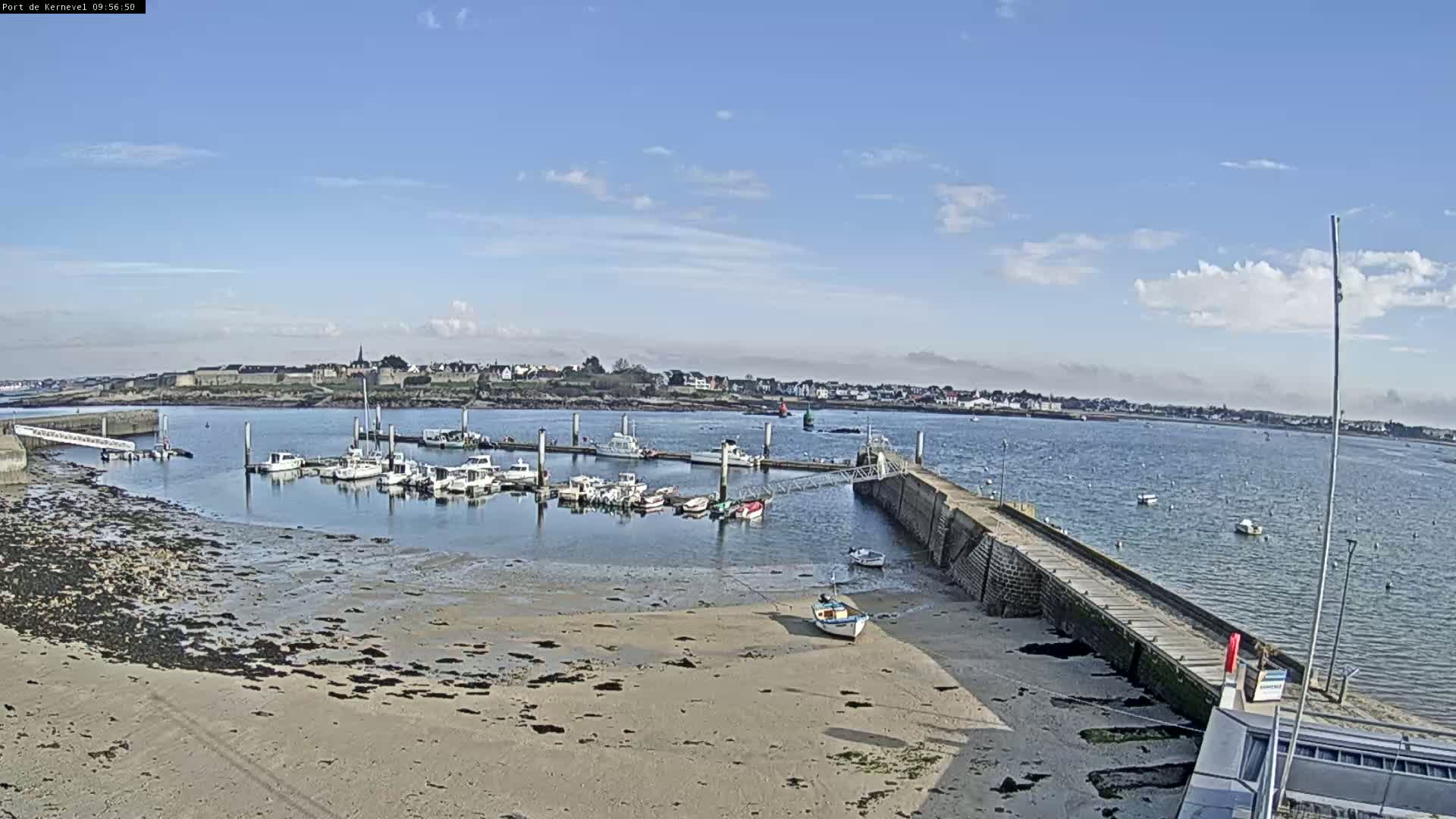 A mostly sunny day shows a harbor with numerous boats docked, a sandy shore, and a small town visible in the distance.
