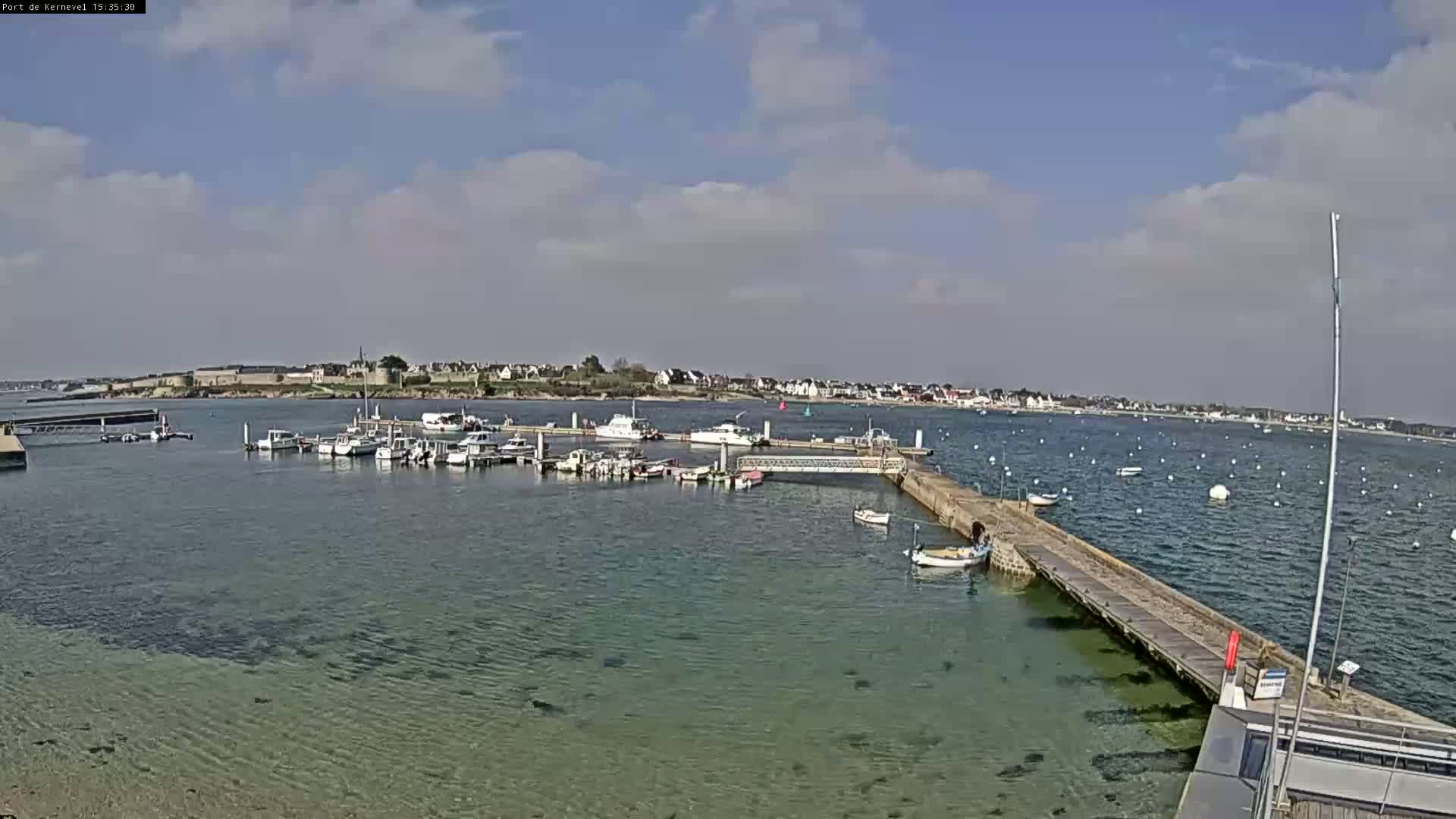 A marina filled with boats sits on calm, clear water under a partly cloudy sky.