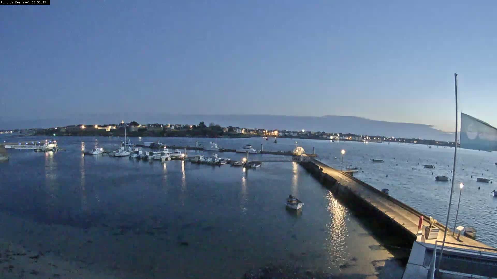 A harbor at twilight is filled with boats moored at a pier extending into calm water, with a town visible in the background.