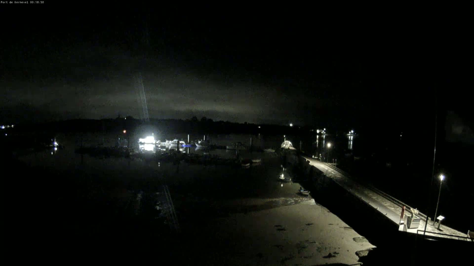 A nighttime view of a harbor with several boats docked, under a mostly cloudy sky.
