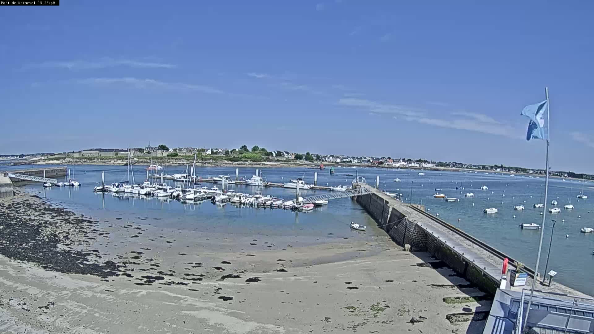 A marina filled with numerous boats is situated on a calm, sunny day next to a sandy shore and a small town in the distance.