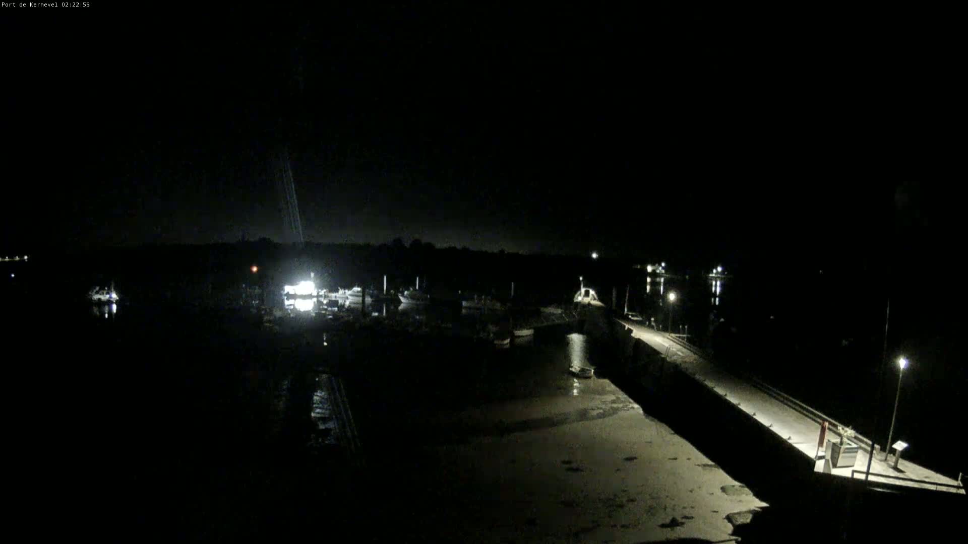 A nighttime view of a harbor with boats docked, a long pier extending into the water, and dimly lit surrounding land under a dark, clear sky.