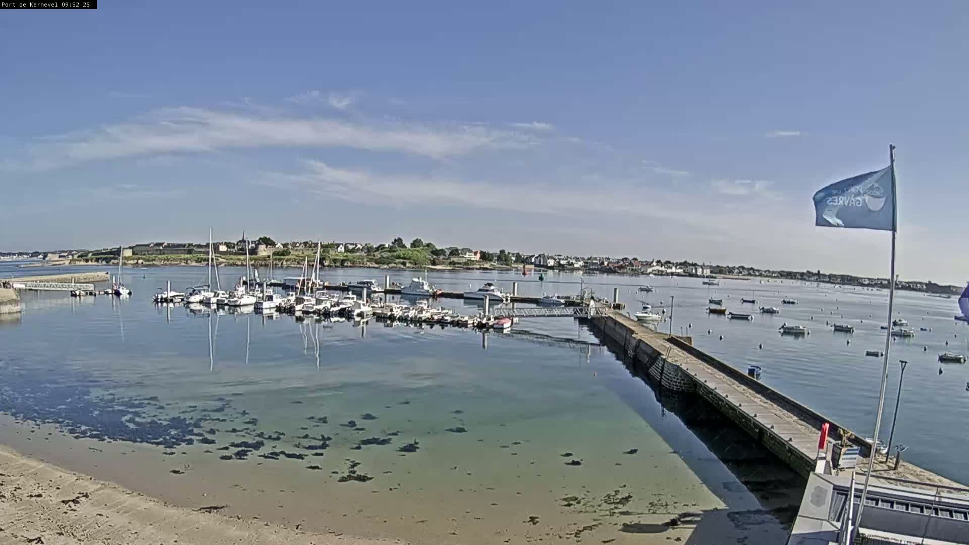 A marina filled with sailboats and motorboats is nestled in a calm bay on a sunny day, with a small sandy beach visible in the foreground.