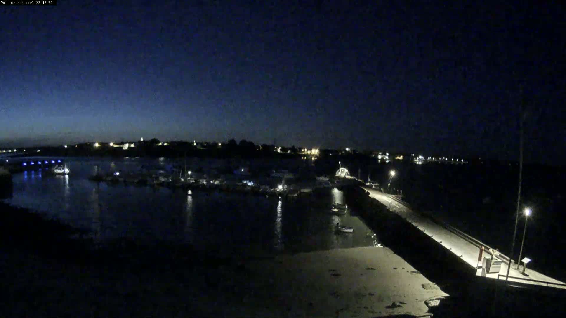 A nighttime view of a harbor filled with boats, a long pier extending into the water, and a town in the distance under a dark sky.