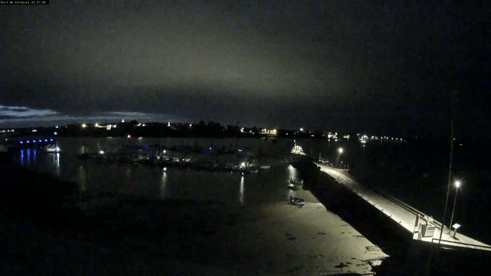 A nighttime view of a harbor filled with boats, a long pier, and a town in the distance under a dark, cloudy sky.