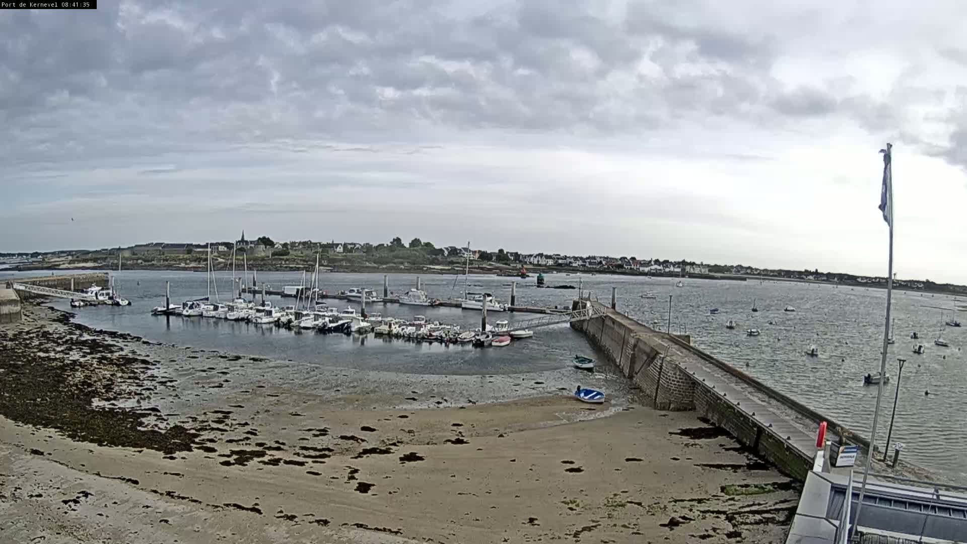 A harbor filled with numerous boats sits under a cloudy sky, with a sandy beach visible in the foreground and additional boats scattered in the water beyond the harbor.