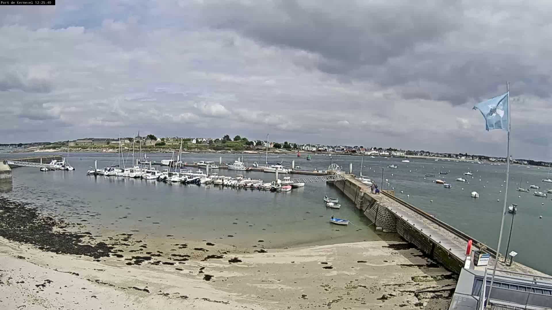 A marina filled with numerous boats sits on a calm body of water under a mostly cloudy sky, next to a sandy beach and a stone pier.