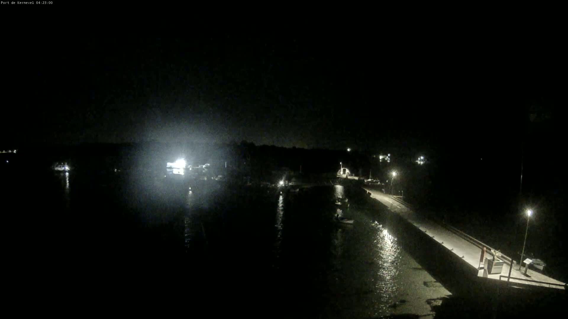 A nighttime view of a dark harbor with a long pier extending into the water, illuminated by several lights along the pier and faint lights from boats and buildings on the shore.