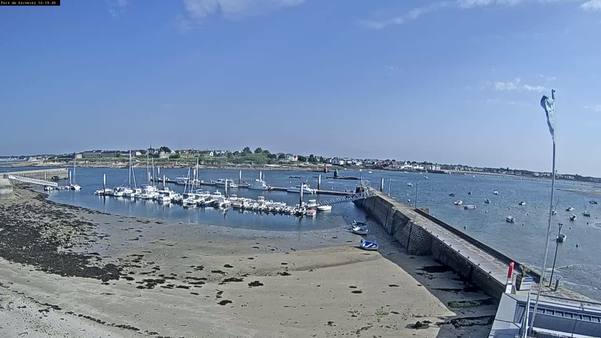 A marina filled with boats is situated on a sandy shoreline, under a clear blue sky, with a small town visible in the background.