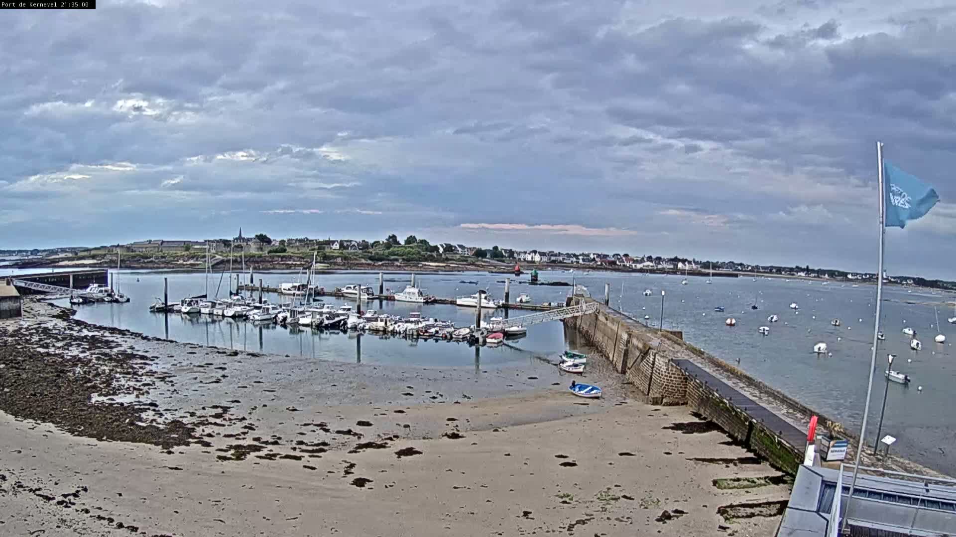 A marina filled with numerous boats is situated on a calm body of water under a mostly cloudy sky, with a sandy shore in the foreground.