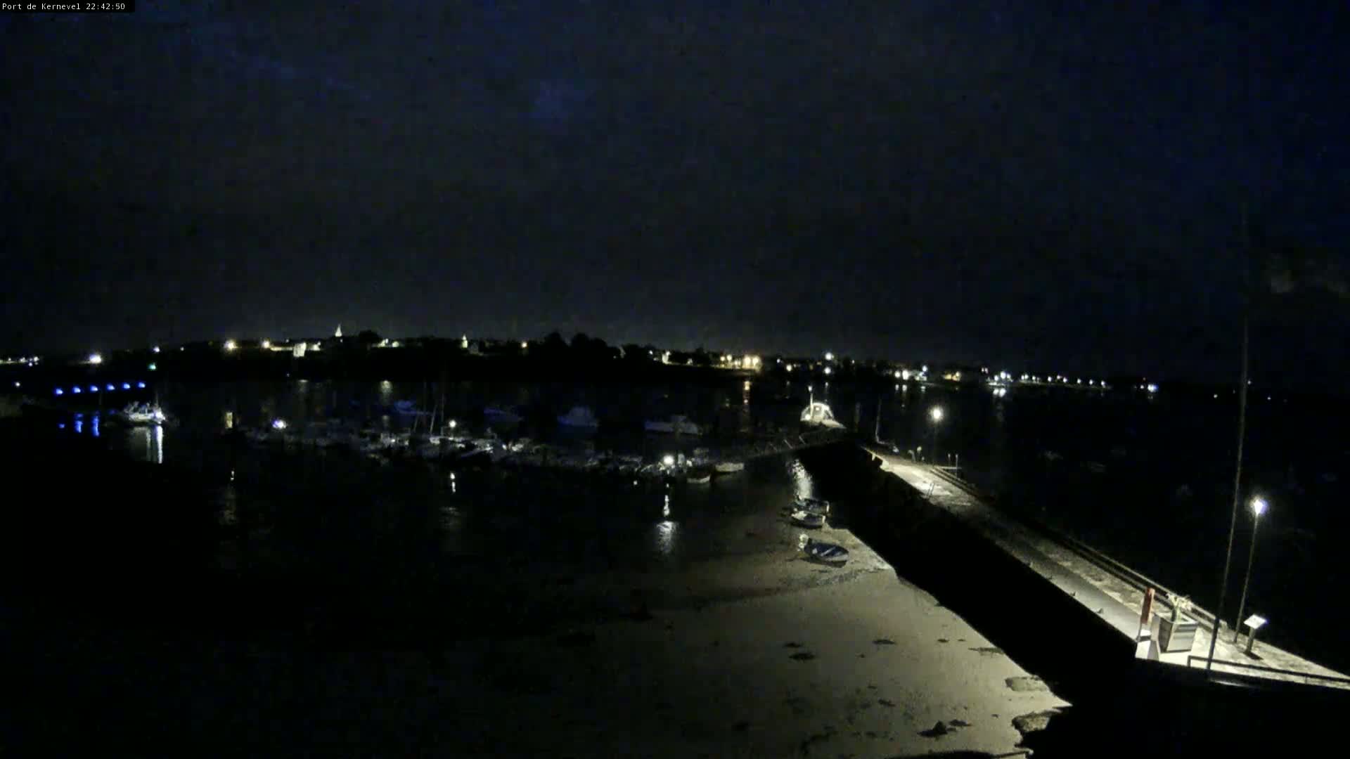 A nighttime view of a harbor filled with boats, a long pier extending into the water, and a dark, overcast sky.