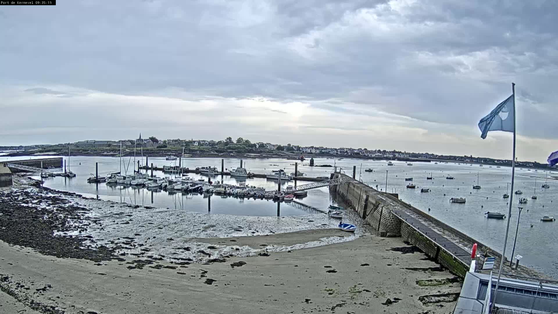 A harbor filled with numerous boats is visible under a cloudy sky, with a sandy shore in the foreground.