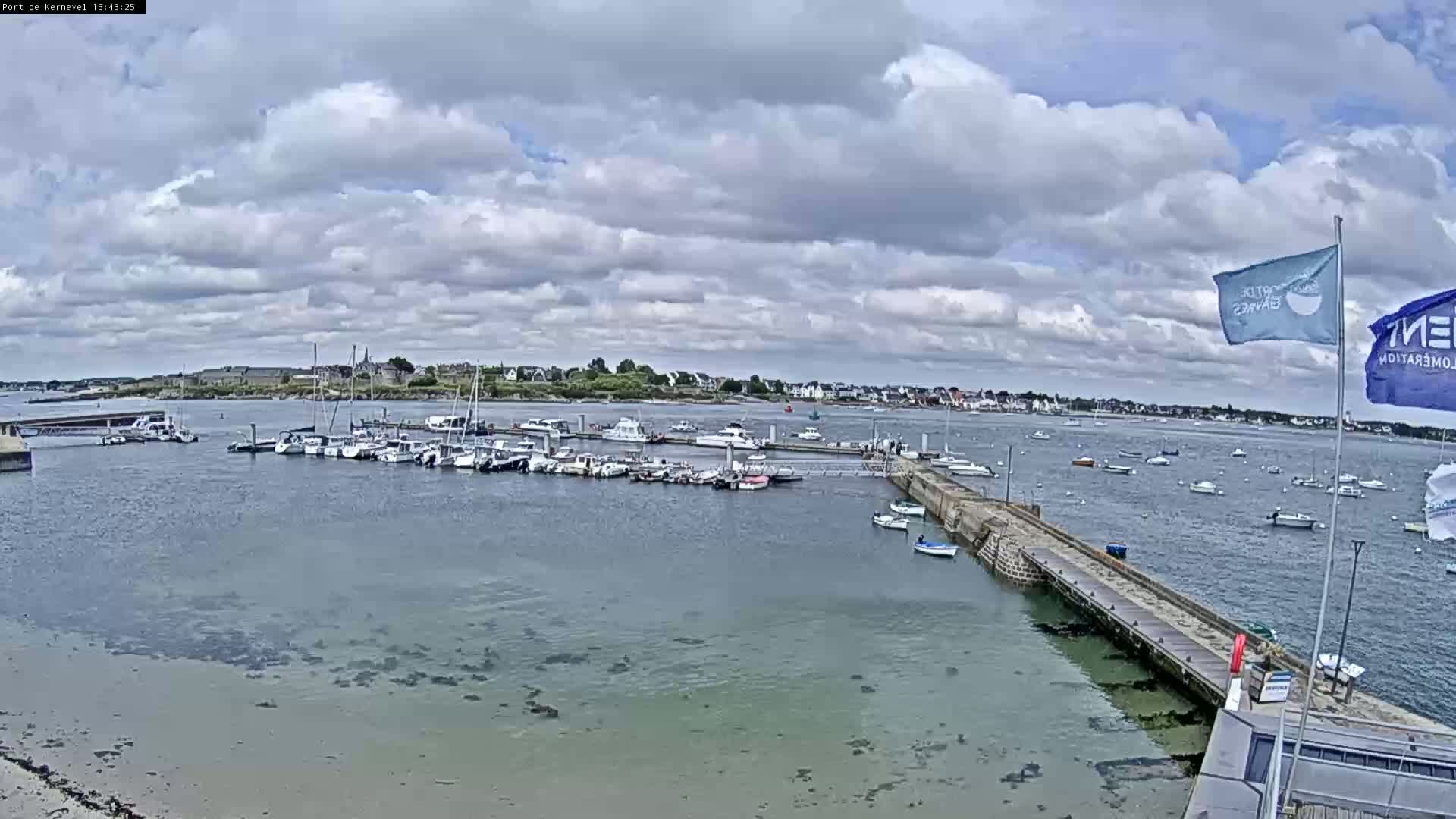 A marina filled with numerous boats is shown under a partly cloudy sky, with calm water and a distant town visible in the background.
