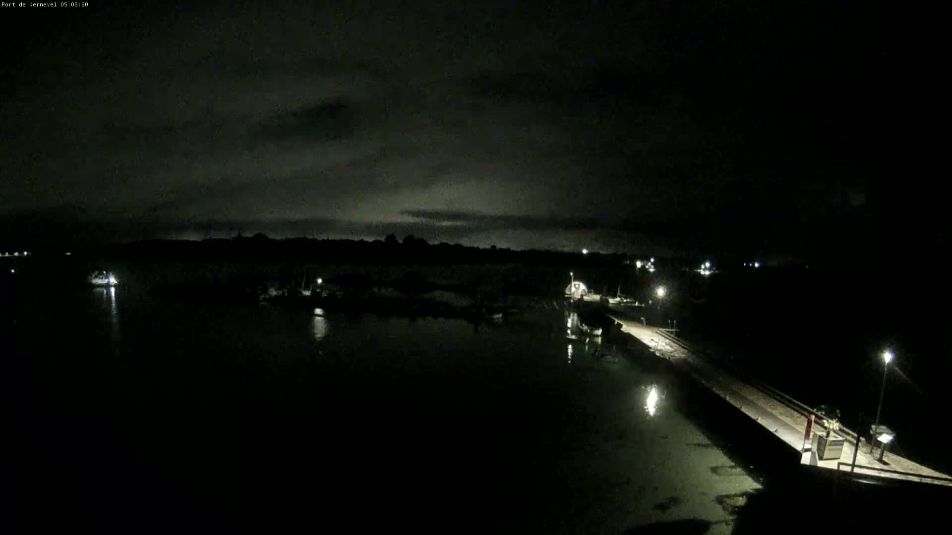 A dark, nighttime harbor scene shows several boats moored in calm water near a lit pier under a cloudy sky.
