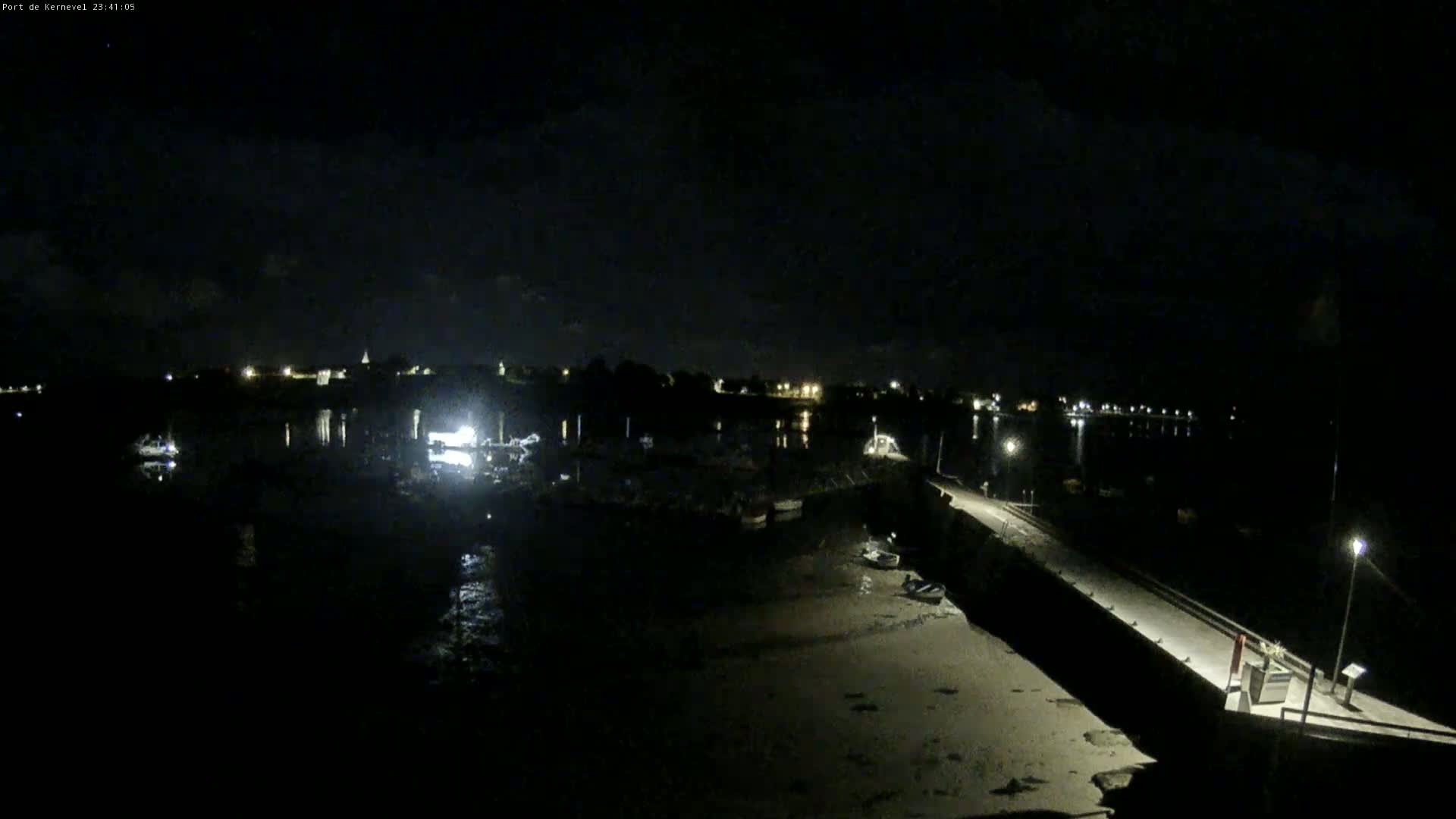 A nighttime view of a harbor with boats docked, a long pier extending into the water, and a town's lights faintly visible in the distance under a partly cloudy sky.