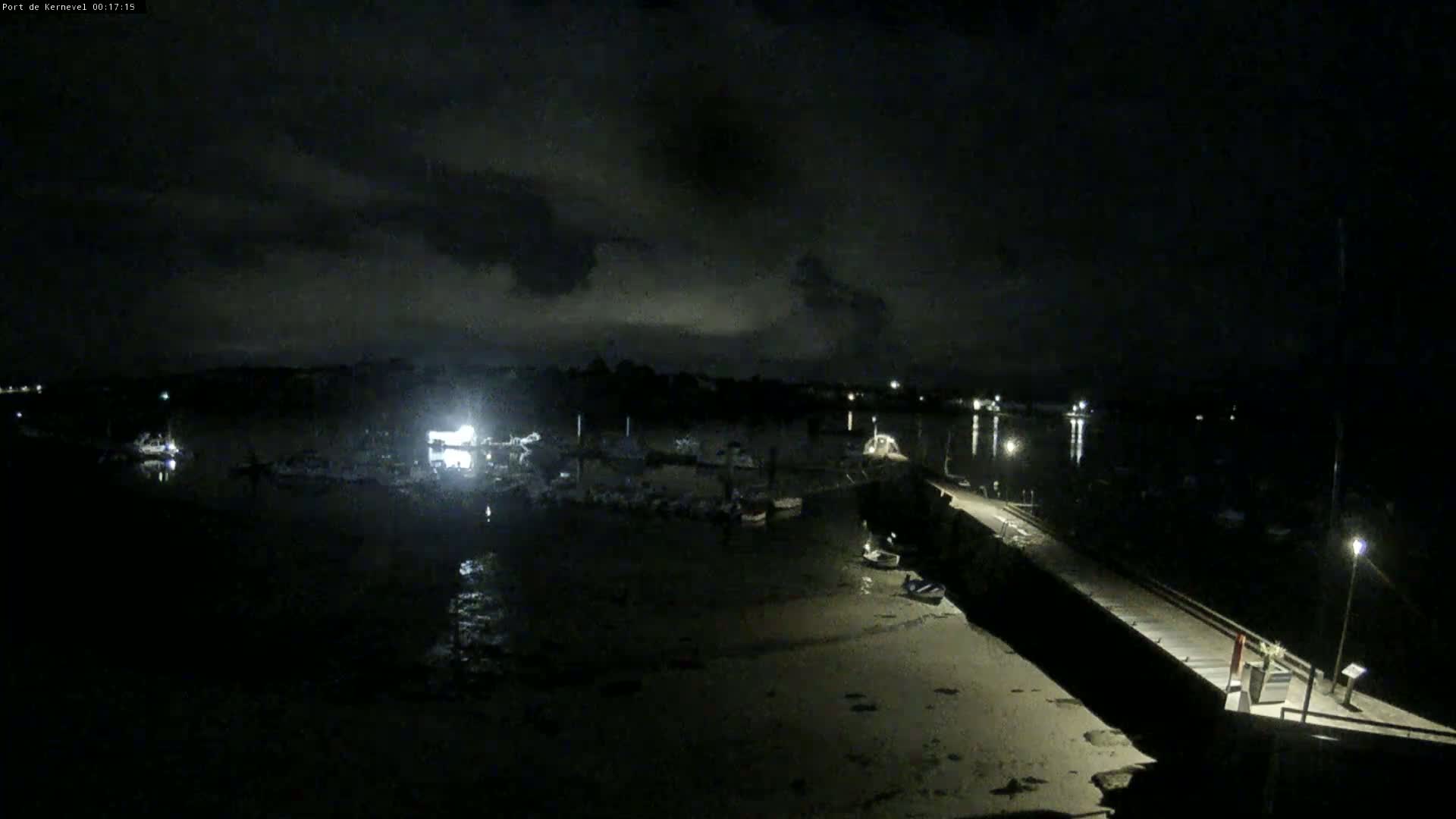 A nighttime, cloudy view of a harbor with boats docked, a pier, and some lights illuminating the scene.