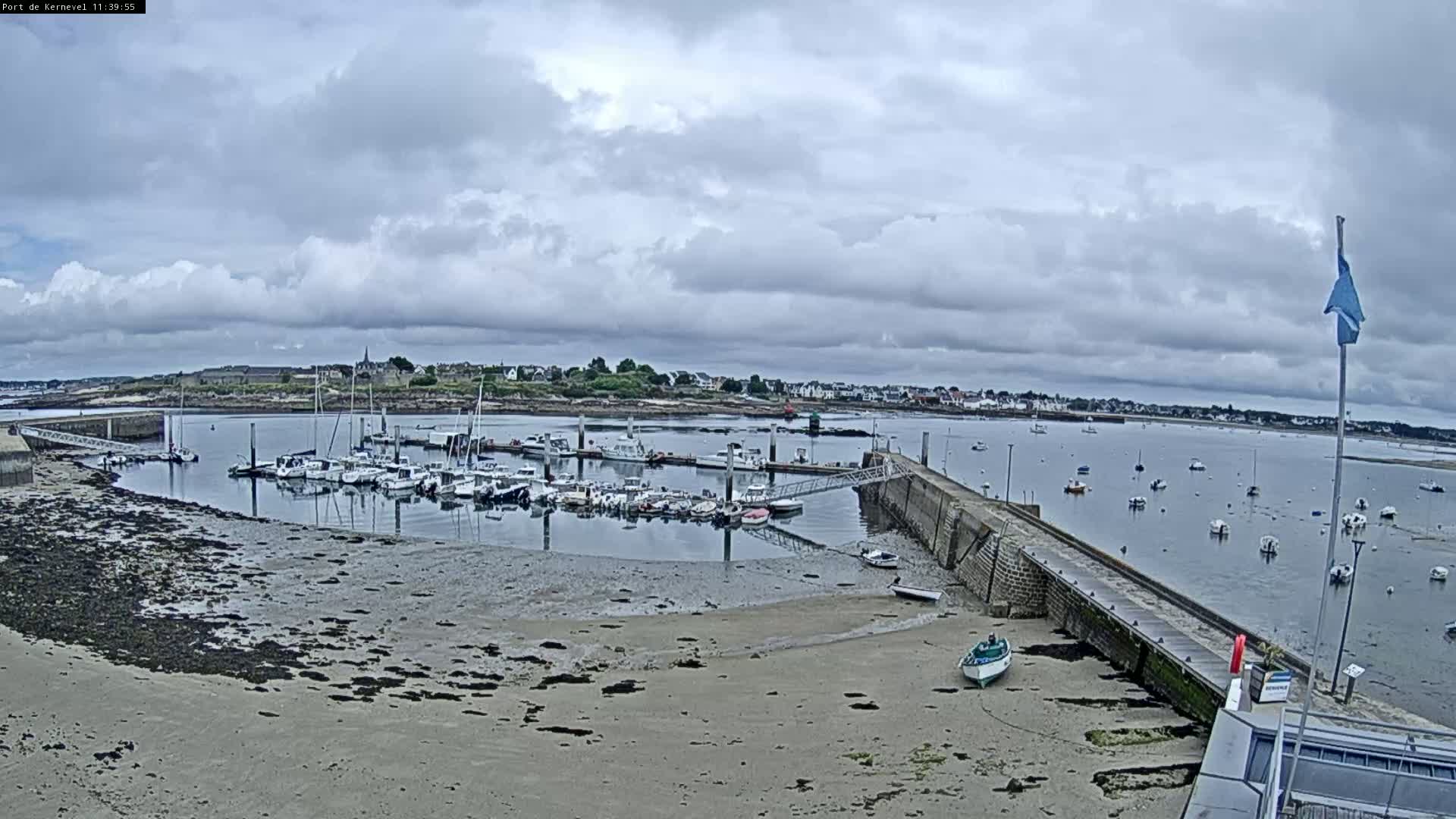 A harbor filled with numerous boats sits under a cloudy sky, with a sandy, low-tide shoreline visible in the foreground.