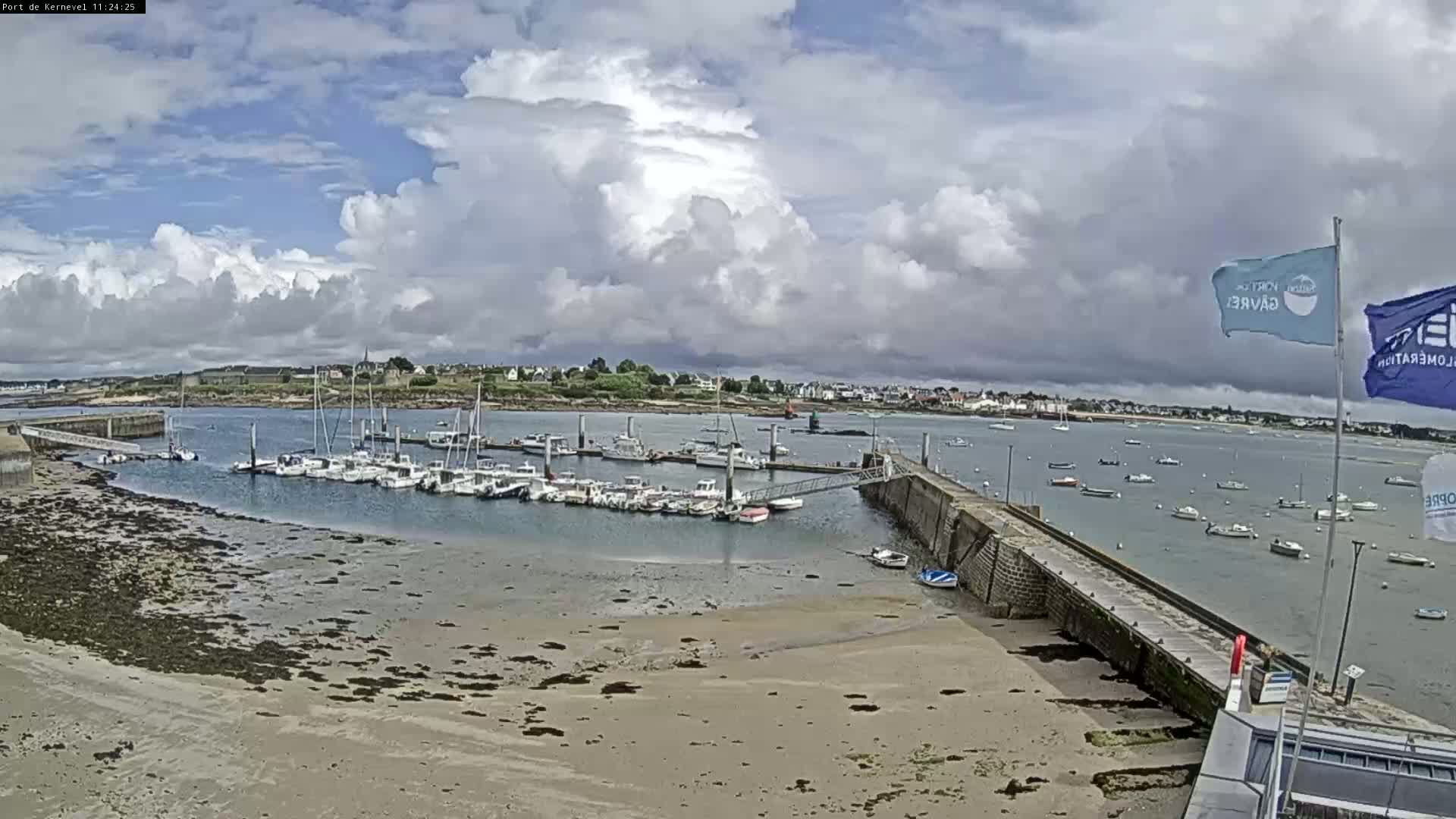 A mostly sunny day with partly cloudy skies shows a harbor filled with sailboats and small motorboats, with a sandy shore and a stone pier in the foreground.