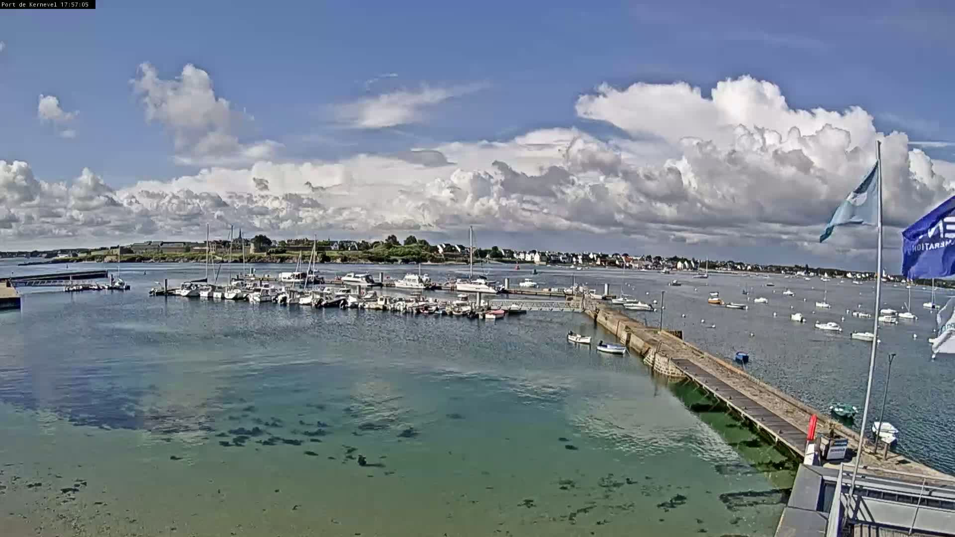 A marina filled with numerous boats is situated in calm, clear water under a partly cloudy sky.