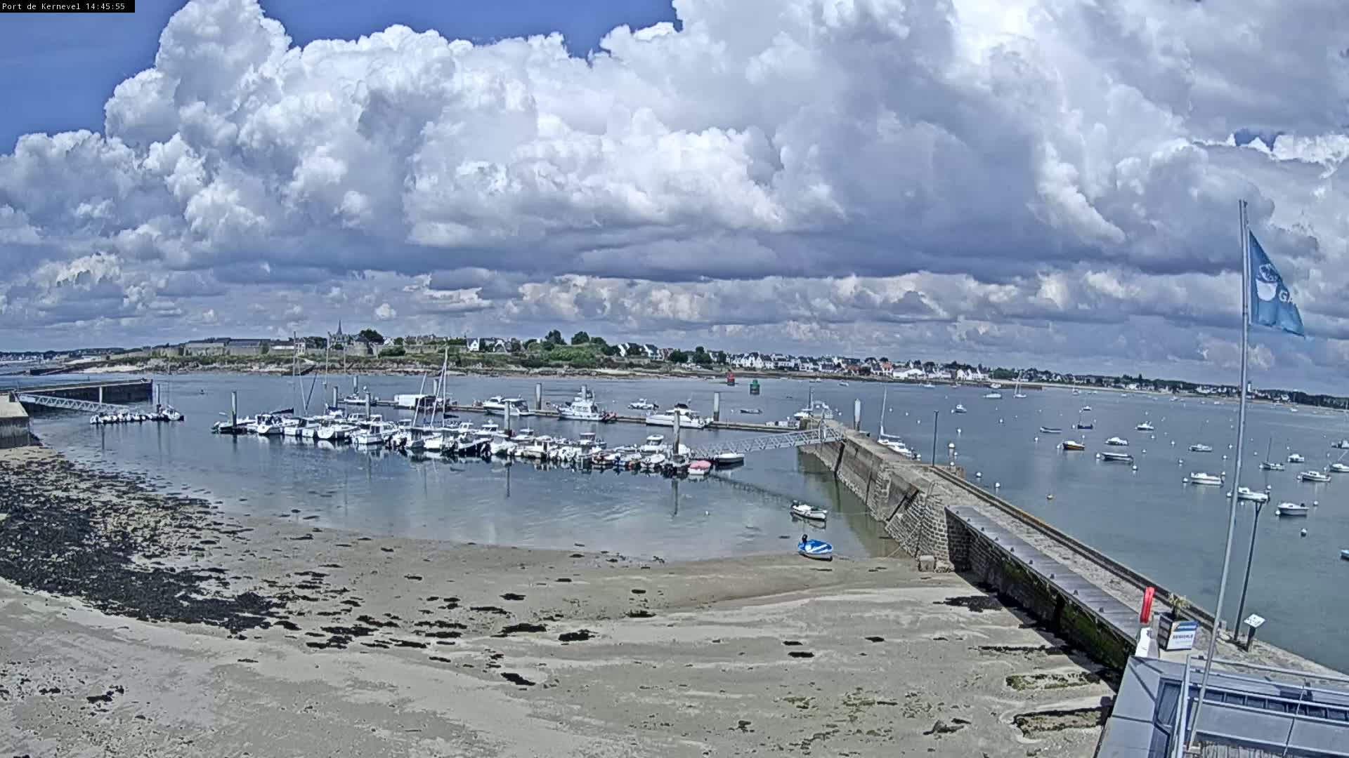 A harbor filled with numerous boats is visible under a partly cloudy sky, with a sandy shore in the foreground.