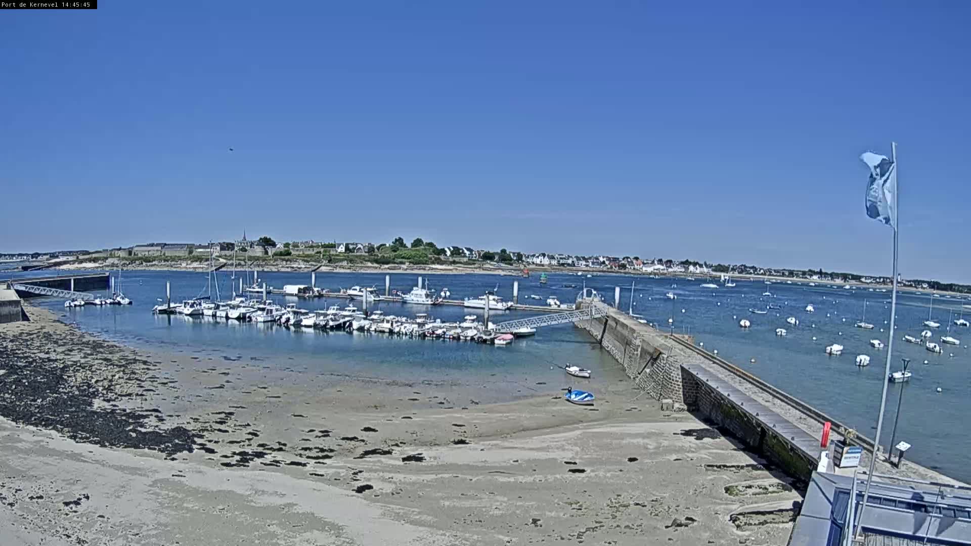 Under a clear blue sky, a marina filled with numerous boats is partially exposed at low tide, with additional boats moored in the calm water beyond a stone jetty.