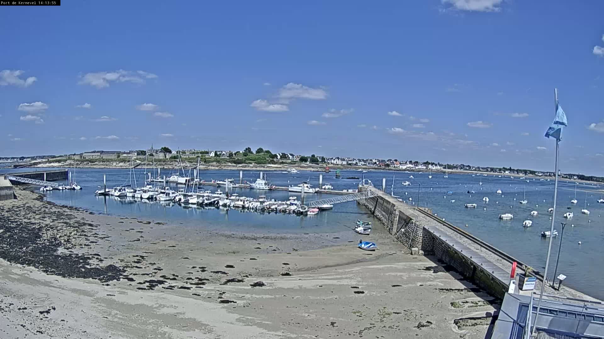 A marina filled with boats sits on a calm, sunny day next to a sandy shore and a stone pier, with a town visible in the background.