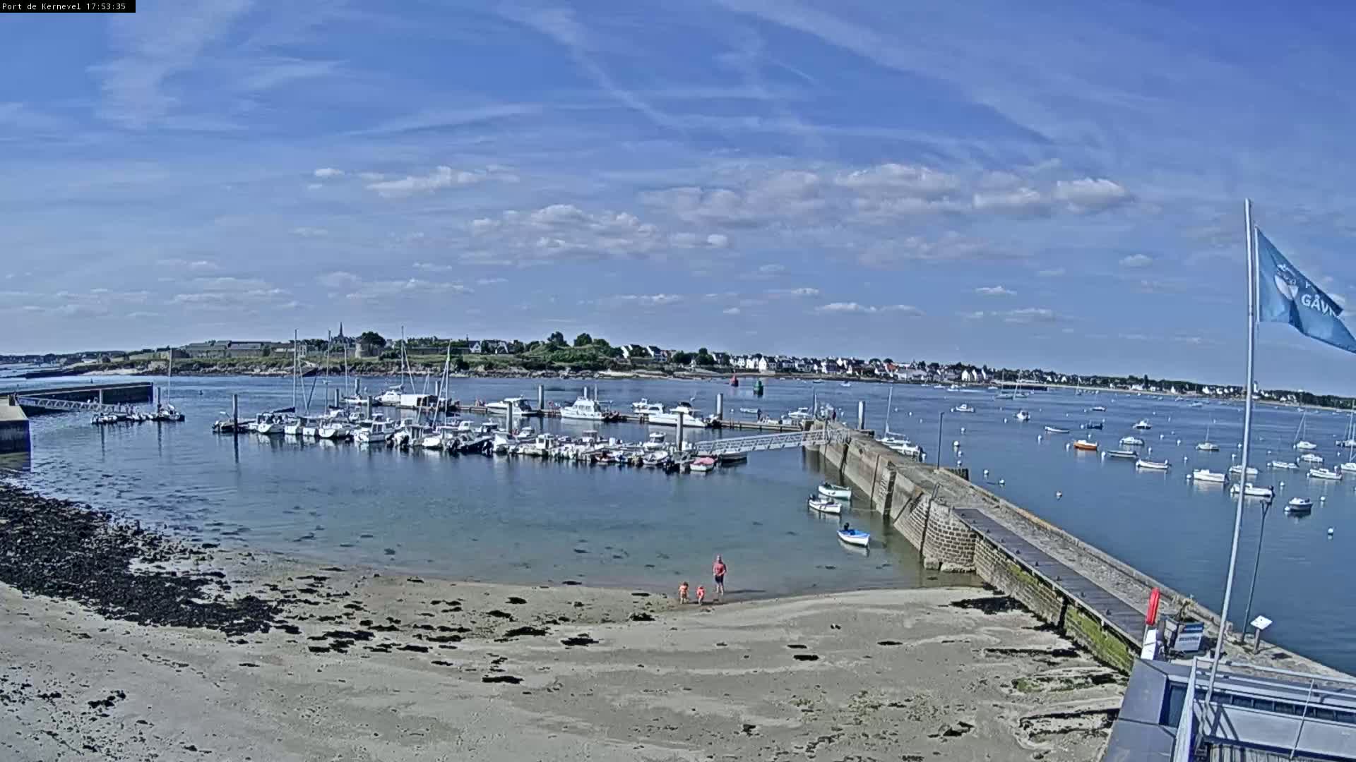 A sandy beach with a few people on it overlooks a calm harbor filled with numerous boats under a mostly sunny sky.