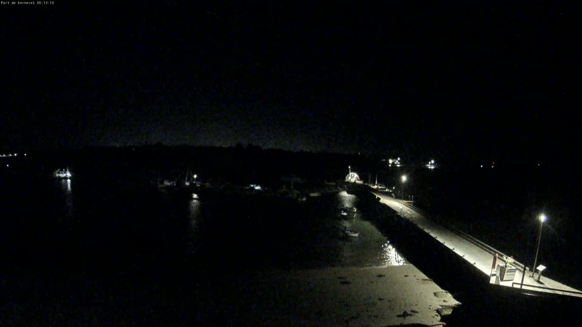 A nighttime view of a harbor with several boats moored, illuminated by faint lights along a stone pier under a dark sky.