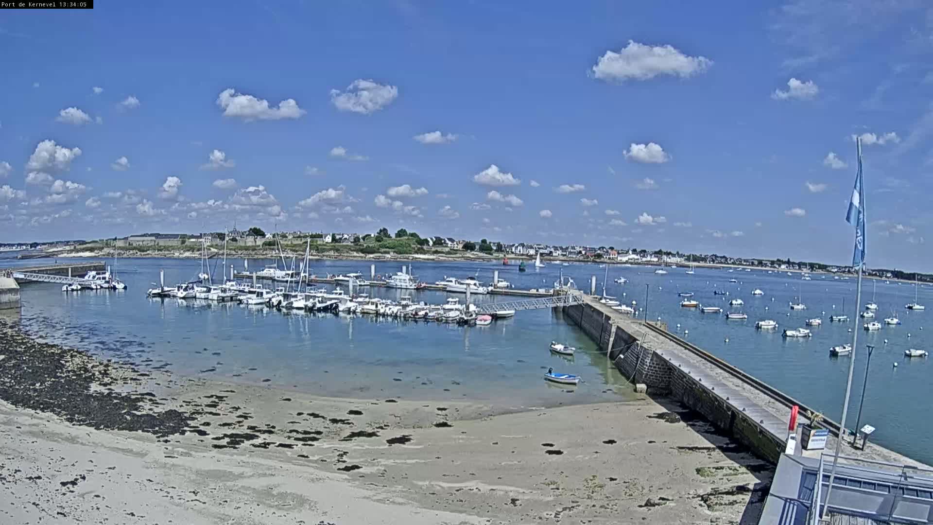 A harbor filled with numerous boats is situated on a sandy shore under a mostly sunny sky with scattered clouds.