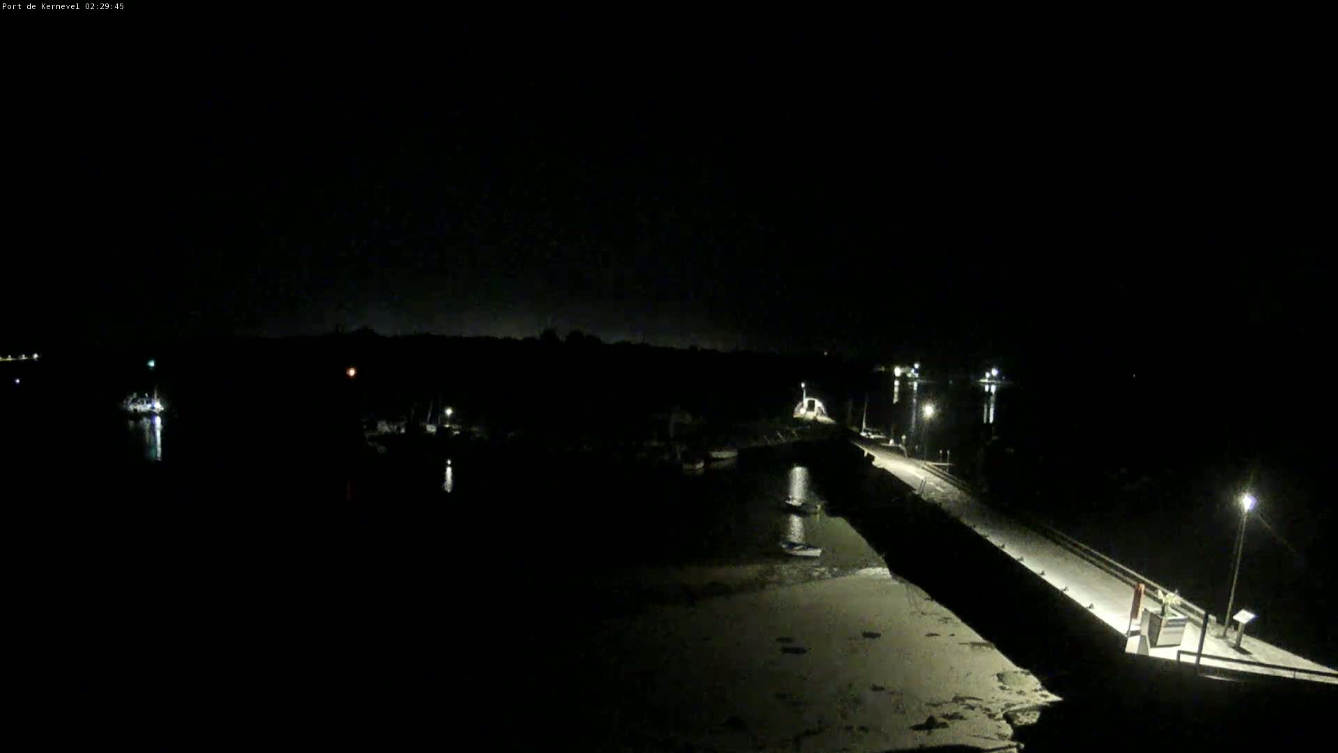 A dark, nighttime view of a harbor with several boats moored, a long pier extending into the water, and a sandy shoreline.