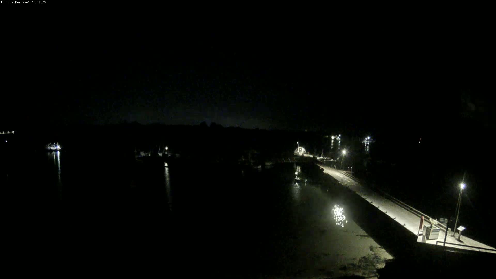 A nighttime, low-light view of a harbor with several boats docked, a long pier extending into the water, and sparsely lit shoreline.
