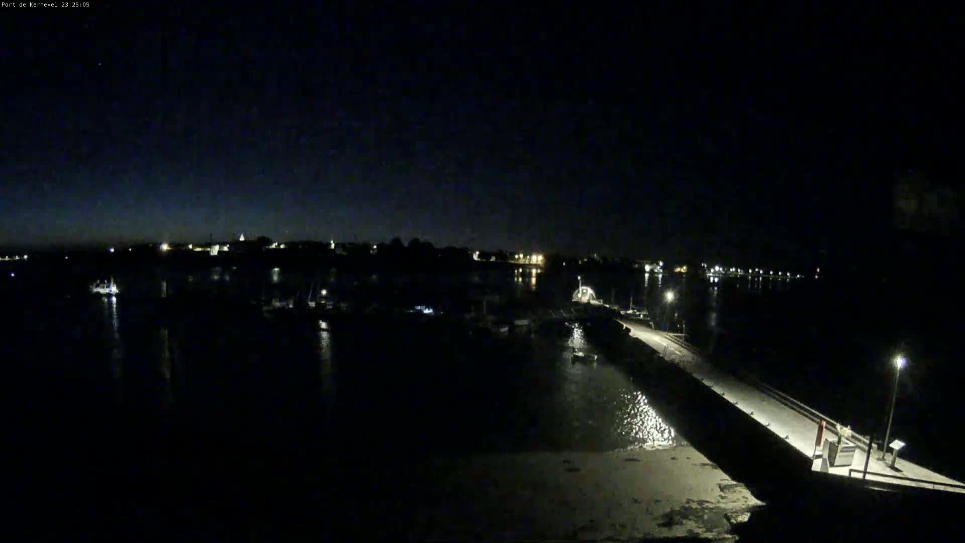 A nighttime view of a harbor with numerous boats docked, a long pier extending into the water, and a dark sky.