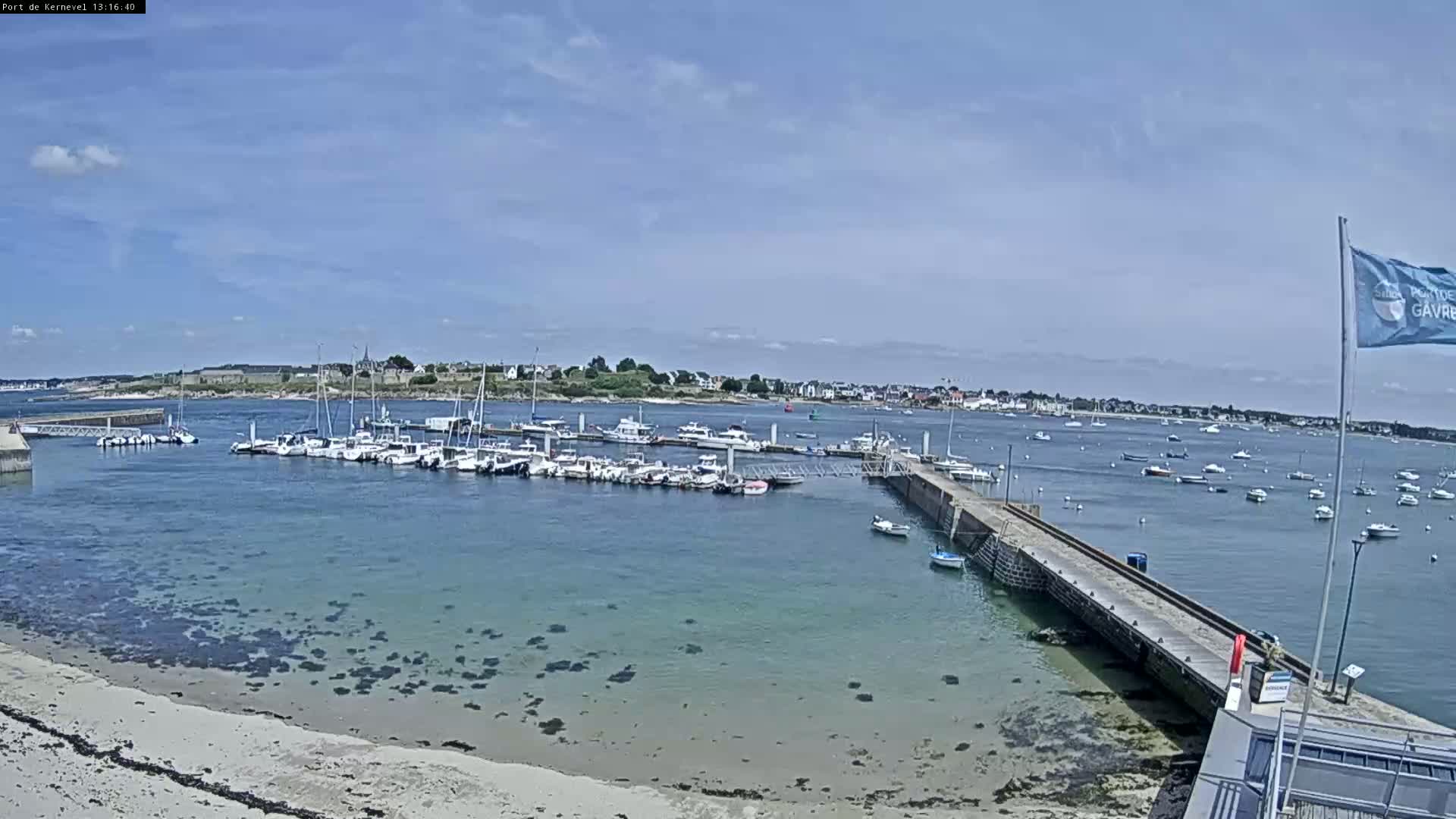 A sunny day shows a harbor filled with sailboats and motorboats, a sandy beach, and a small town in the distance.