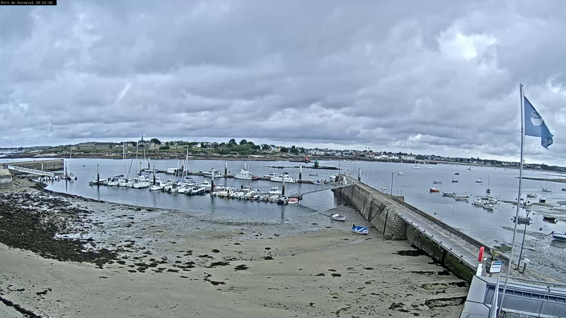 A harbor filled with numerous boats under a cloudy sky, with a sandy shore and a low-lying pier extending into the water.