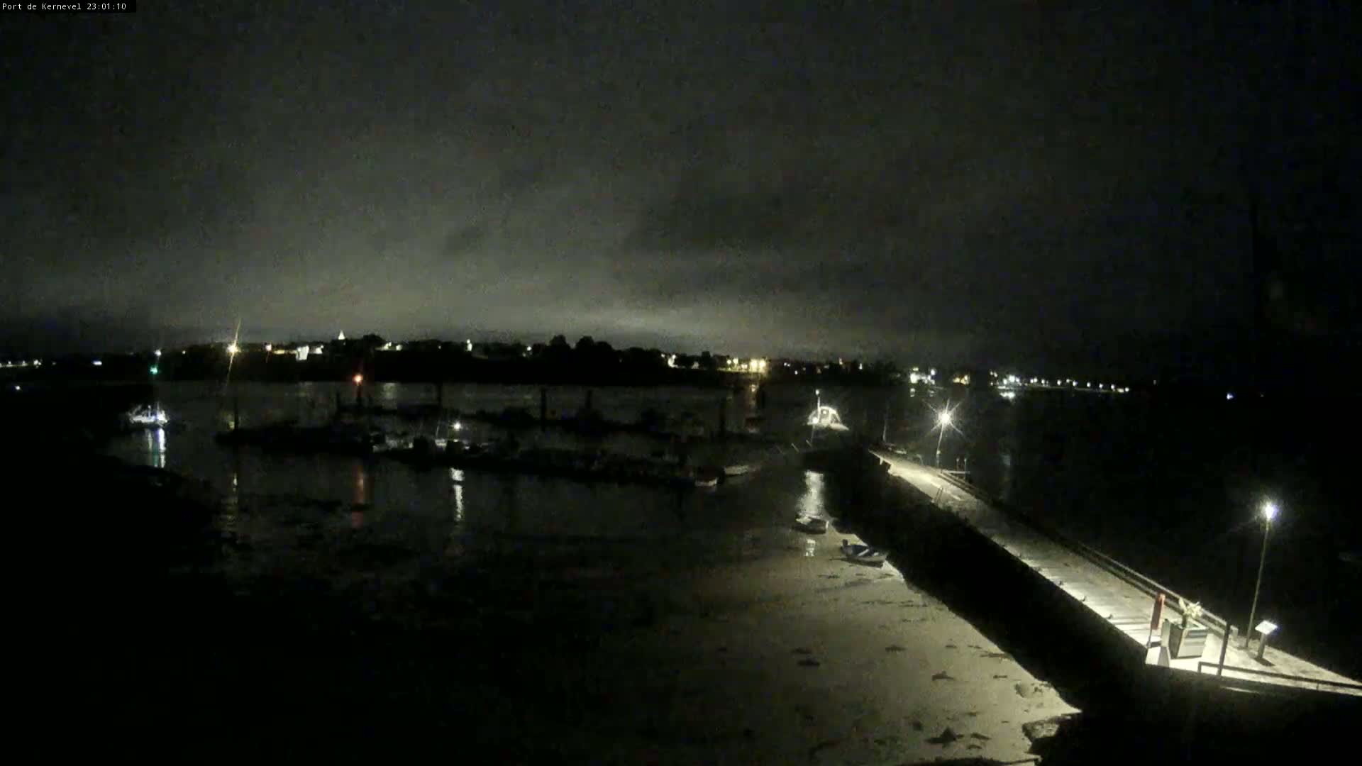 A nighttime view of a harbor with several boats moored, under a dark, cloudy sky.
