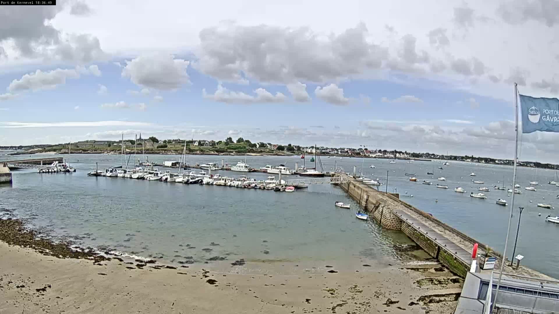 A partly cloudy day reveals a harbor filled with numerous boats, a sandy shore, and a small town in the distance.