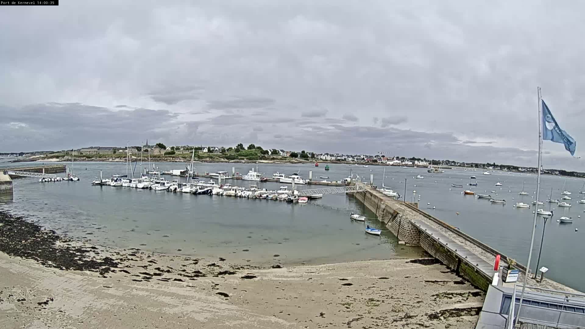 A harbor filled with numerous boats is situated near a sandy beach under an overcast sky.
