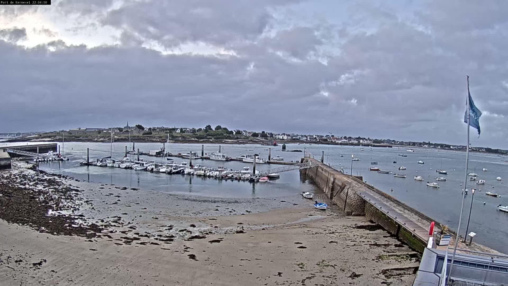 A harbor filled with many small boats sits under a cloudy sky, with a sandy shore and a stone pier extending into the water.