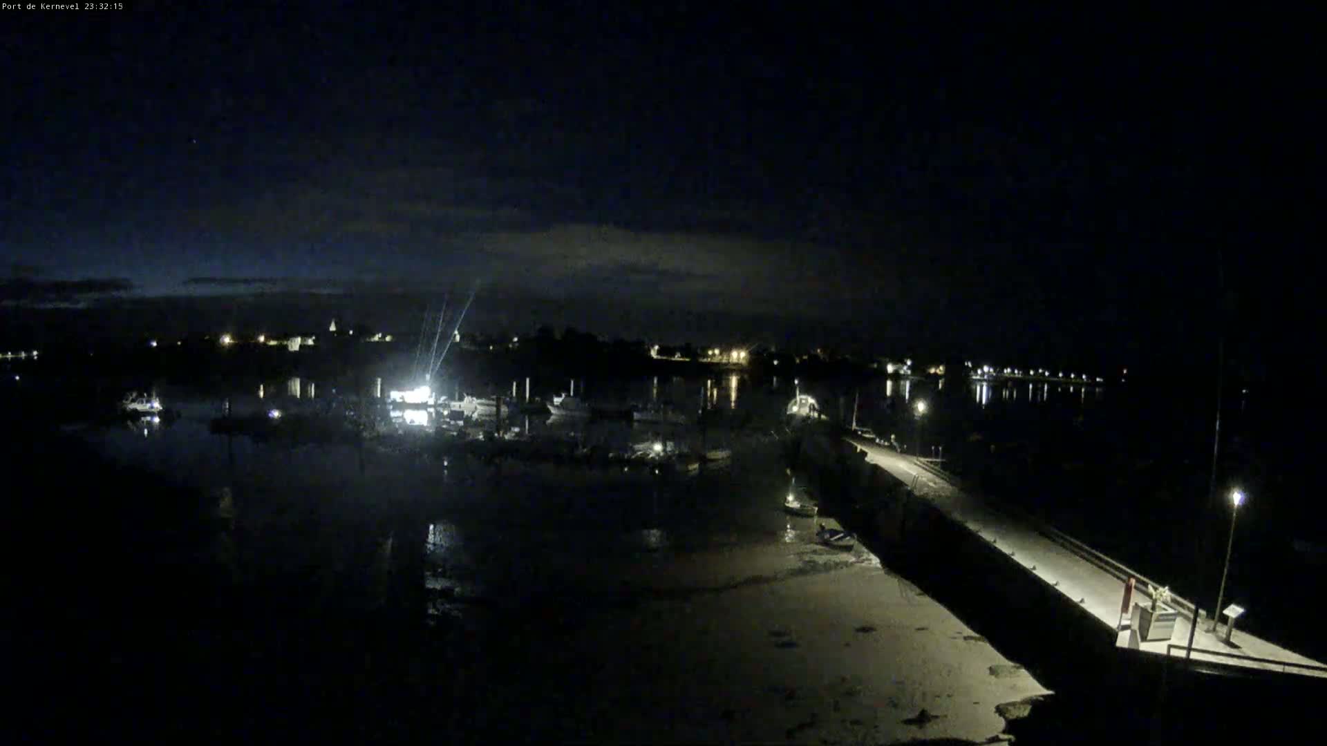 A nighttime view of a harbor with boats moored, a long pier extending into the water, and a town faintly visible in the distance under a mostly clear sky.
