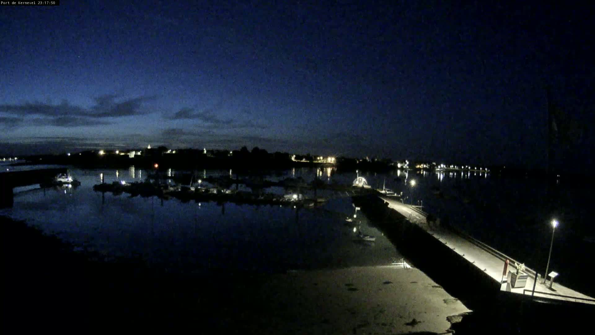 A nighttime harbor scene shows a calm body of water with docked boats reflecting the lights of a nearby town under a dark, mostly clear sky.