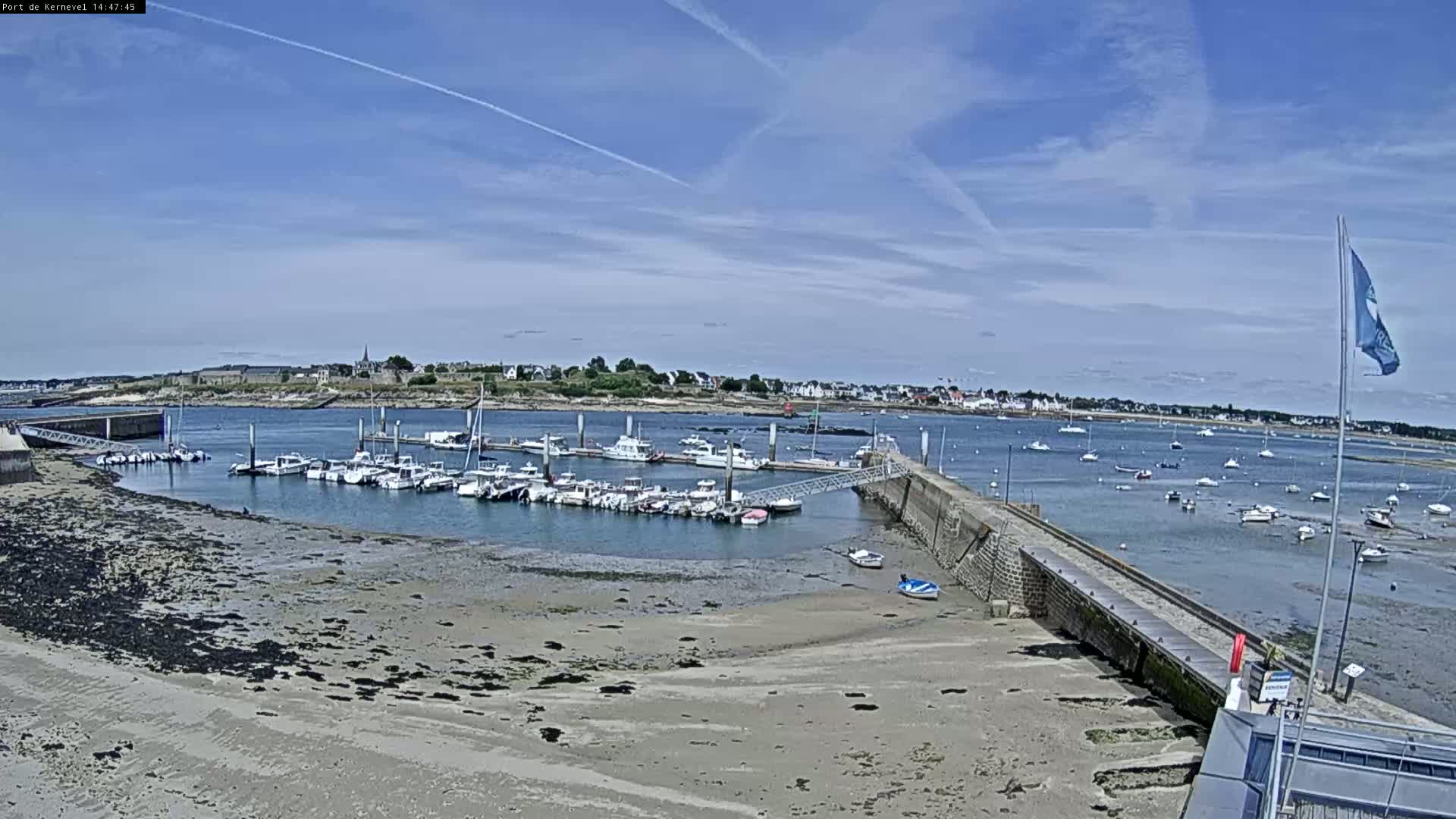 A marina filled with numerous boats is situated on a sandy shore under a clear blue sky with a few contrails.