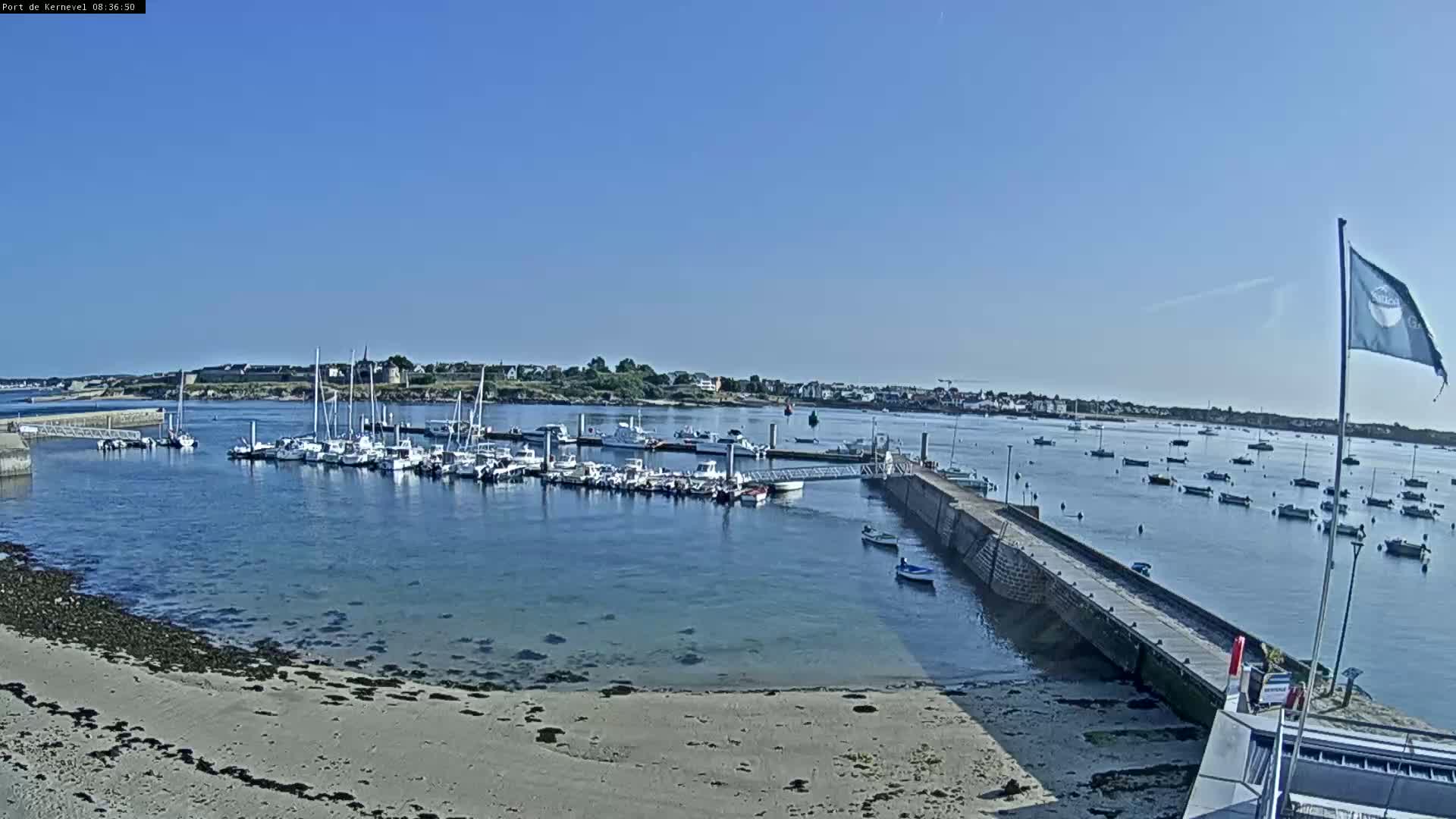 A sunny day at a harbor shows many boats docked and others anchored in the water, with a sandy beach in the foreground.