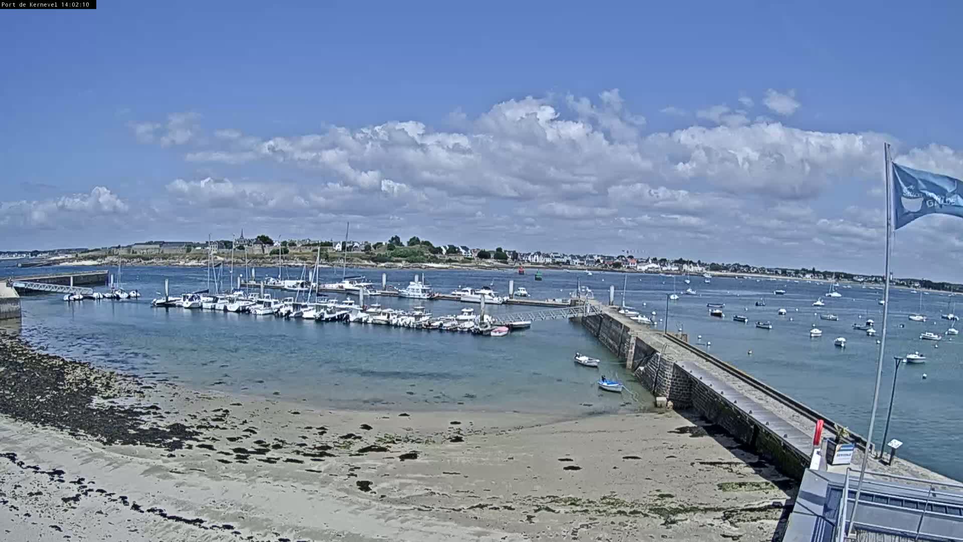 A mostly sunny day shows a harbor filled with many boats, a sandy beach, and a small town in the distance.