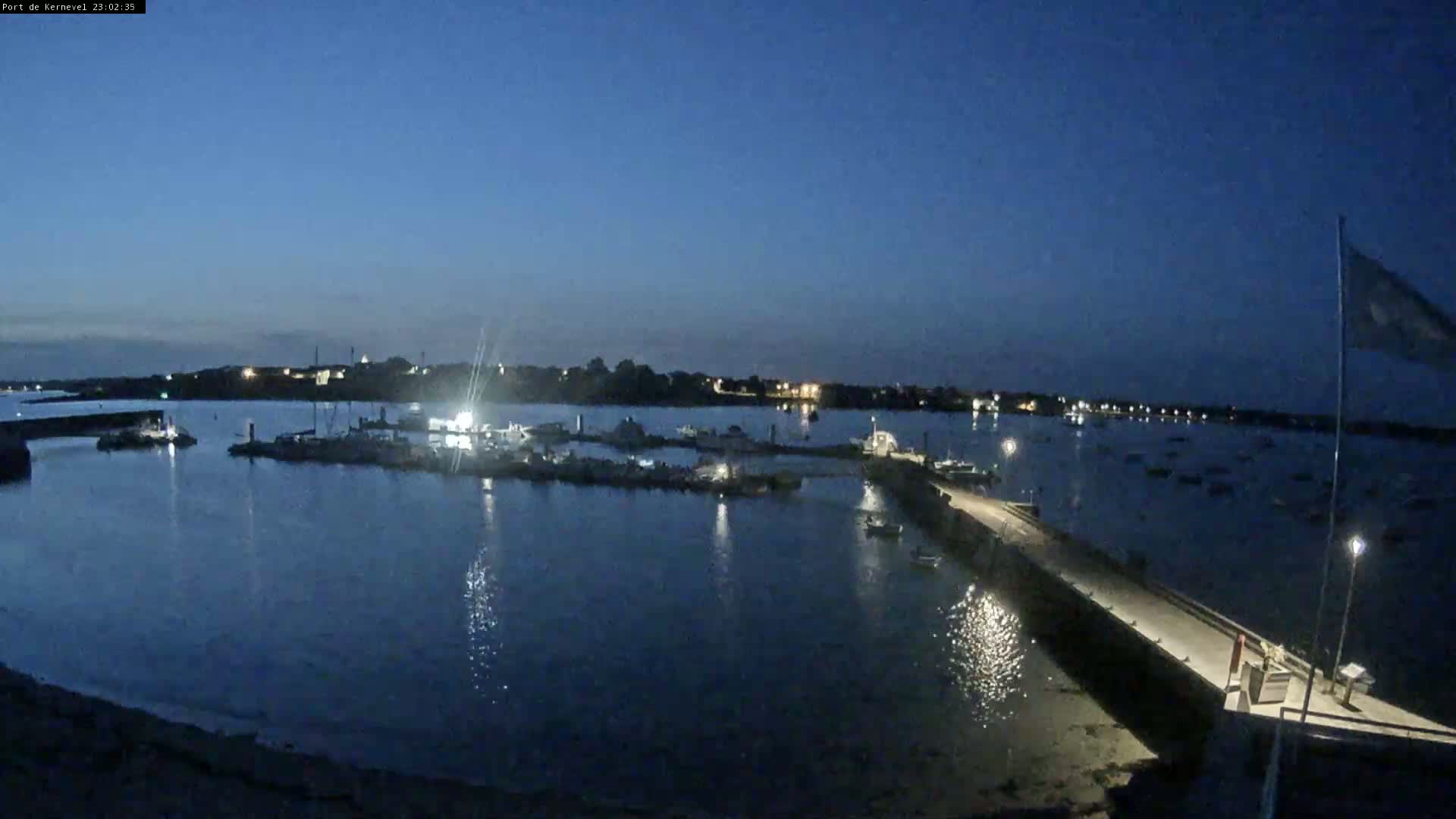A nighttime view of a calm harbor with numerous boats docked, illuminated by faint lights under a dark blue sky.