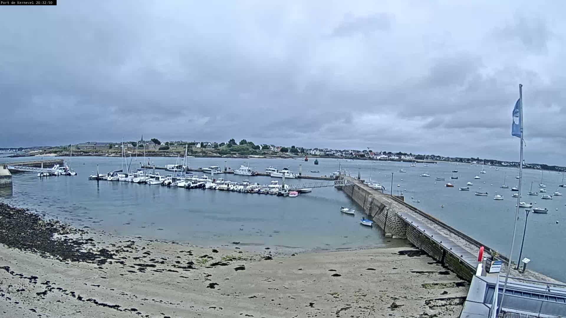 A harbor filled with numerous boats is seen on an overcast day, with a sandy beach and a stone pier extending into the water.