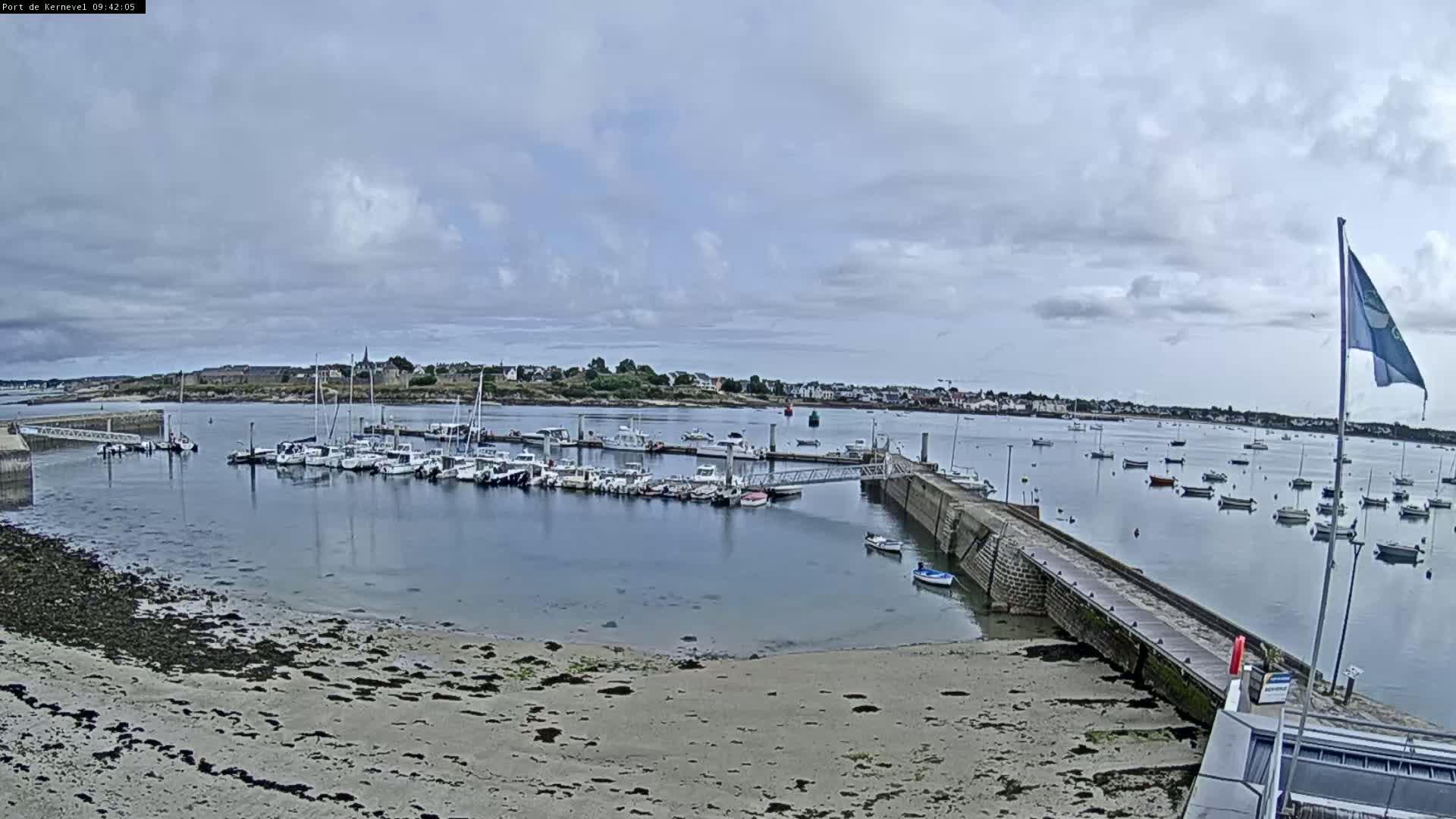 A harbor filled with numerous boats under a mostly cloudy sky, with a sandy shore in the foreground.