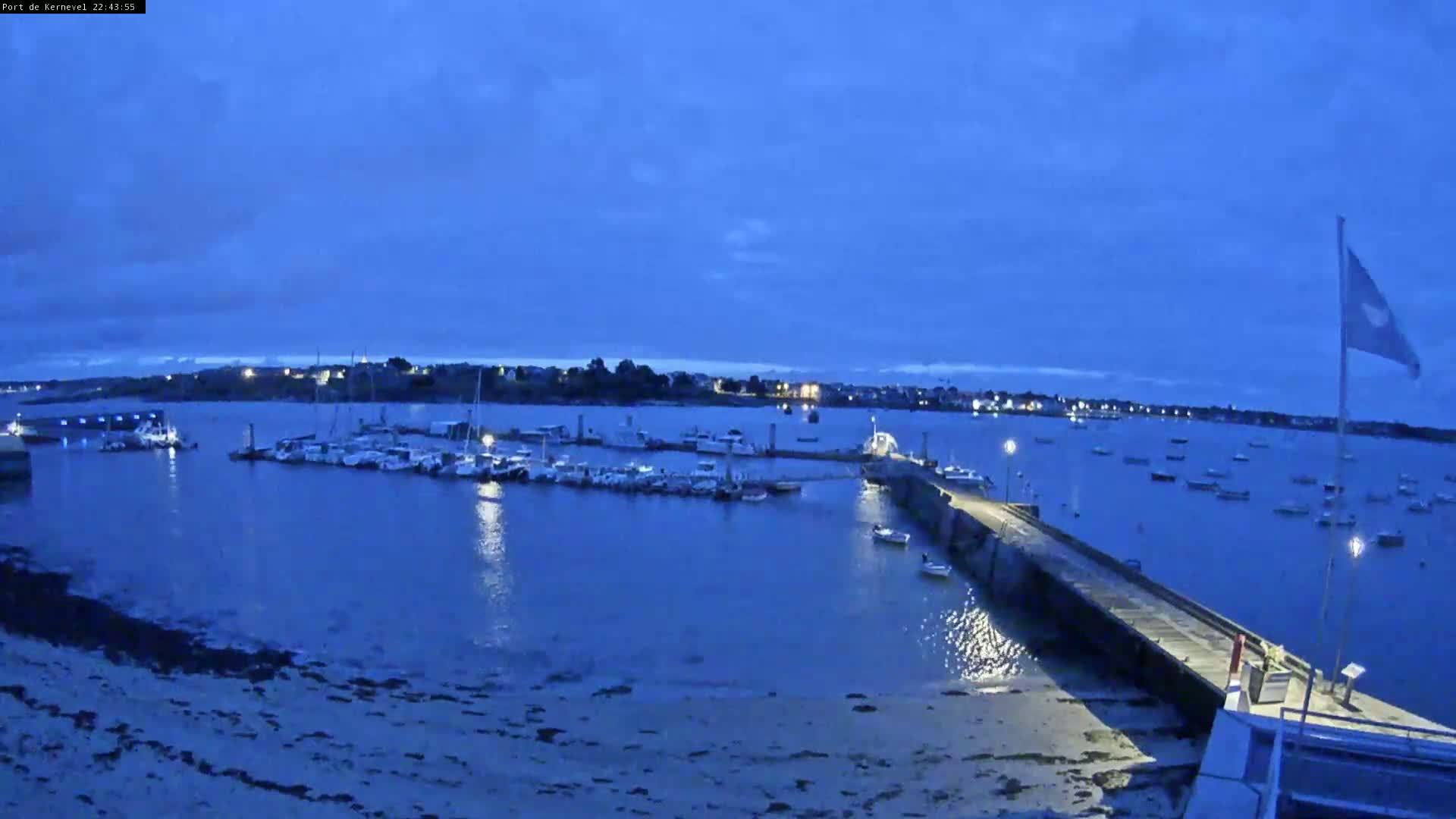 Under a twilight blue sky, a marina filled with boats is seen from a shore with a long pier extending into the water.